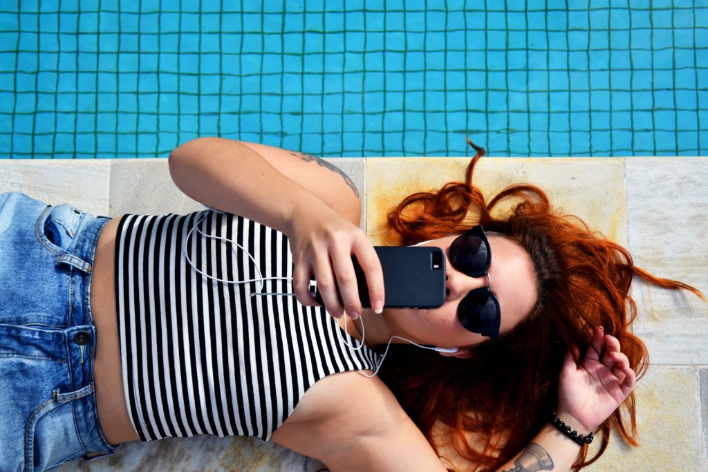 Latin woman lying by the pool with sunglasses and tanktop on while talking to her phone