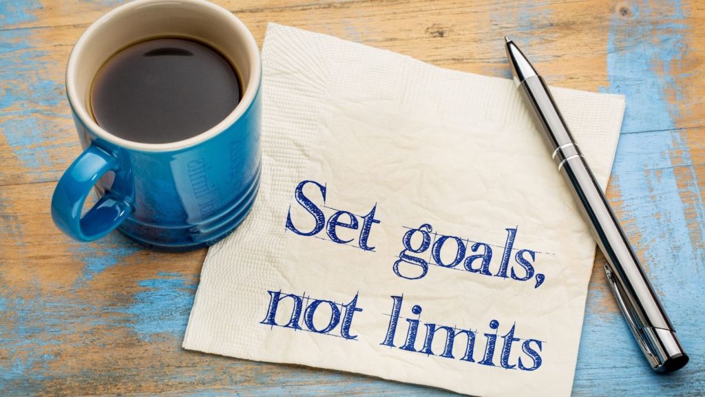 a blue coffee mug with coffee to the left sitting on a wooden table with a pen opposite and a napkin between with the words set goals, not limits